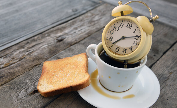 Alarm clock inside a coffee cup with toast on wooden table.