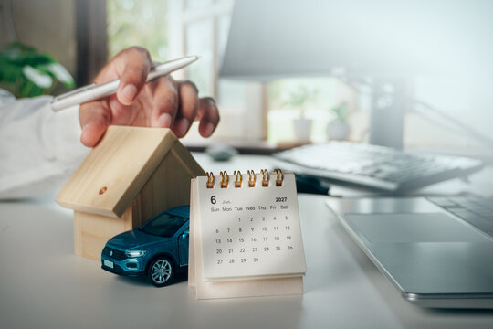 Hand holding a pen over a wooden house model with a toy car and June 2027 calendar on office desk, concept of mortgage, home loan, property and vehicle financial planning.