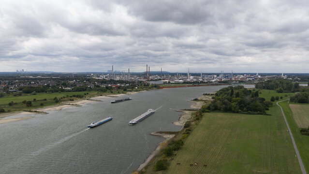 Aerial view of the Dormagen-Bayerwerk industrial complex along the Rhine river with cargo barges navigating the waterway, near green fields in Cologne, North Rhine-Westphalia, Germany.