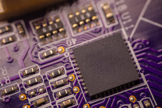Close-up view of a purple printed circuit board featuring a central black microchip and various electronic components, showcasing intricate connections and soldering details