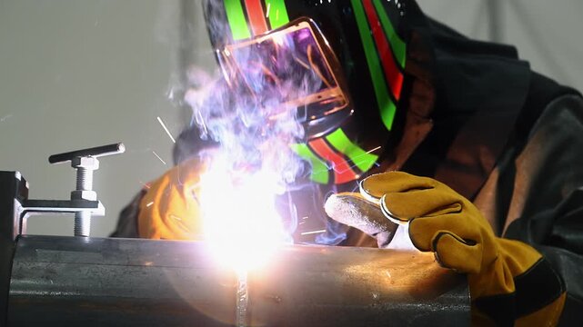 Professional welder in protective mask and gloves welding metal. Bright welding arc, smoke and weld seam show an industrial metal fabrication process.