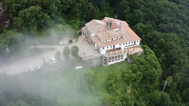 Aerial view of Mare de deu de la salut Hermitage, dramatically perched on a rocky cliff above dense forests, overlooking the beautiful mountain landscapes of Girona, Catalonia. 