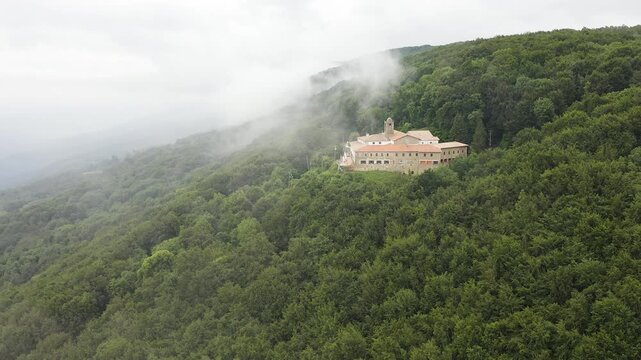 Aerial view of Mare de deu de la salut Hermitage, dramatically perched on a rocky cliff above dense forests, overlooking the beautiful mountain landscapes of Girona, Catalonia. 