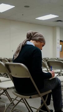 Vertical video: Writing woman in tan hijab taking notes in class with blue pen, pad, folding chairs