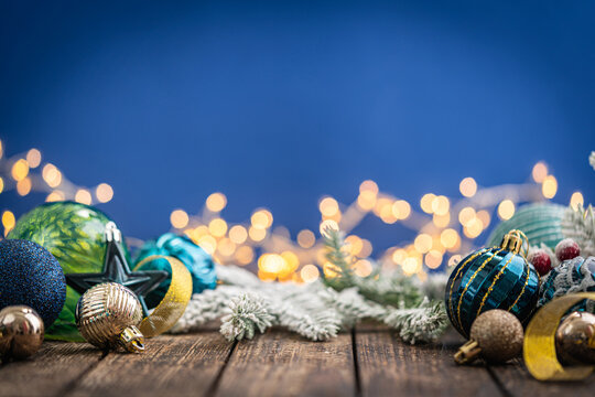 A Christmas background featuring navy and teal ornaments wrapped with gold ribbon on rustic wood, framed symmetrically against a deep blue backdrop with twinkling lights and copy space.