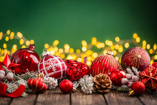 A Christmas background featuring an abundant arrangement of red baubles, a glittering star, and a pinecone nestled in frosted pine on rustic wood, set against green with twinkling lights.