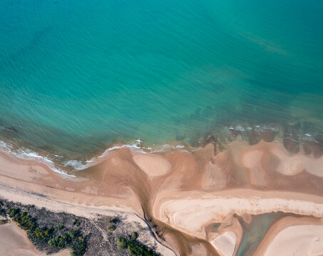 Aerial view of a sandy beach meeting turquoise ocean waters with intricate tidal patterns and coastal vegetation in Australia.