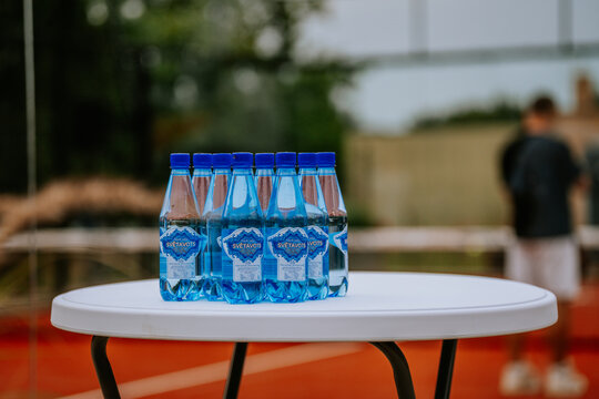 Bottles of blue mineral water arranged on a white table at a padel court, with blurred players and a red clay surface visible in the background