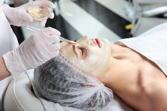 Esthetician spreads cosmetic paste over relaxed female cheeks using brush. Fingers in protective wear hold glass jar. Quiet spa environment