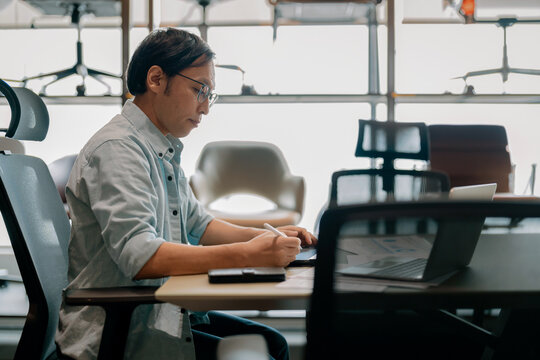 Focused Asian architect drawing on digital tablet with stylus at office desk under warm ambient light, demonstrating concentrated diligence, creative drafting precision, thoughtful planning, absort