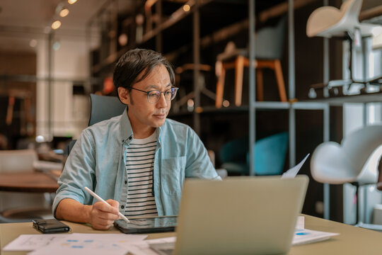 Focused Asian architect drawing on digital tablet with stylus at office desk under warm ambient light, demonstrating concentrated diligence, creative drafting precision, thoughtful planning, absort