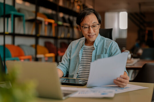 Focused Asian architect drawing on digital tablet with stylus at office desk under warm ambient light, demonstrating concentrated diligence, creative drafting precision, thoughtful planning, absort