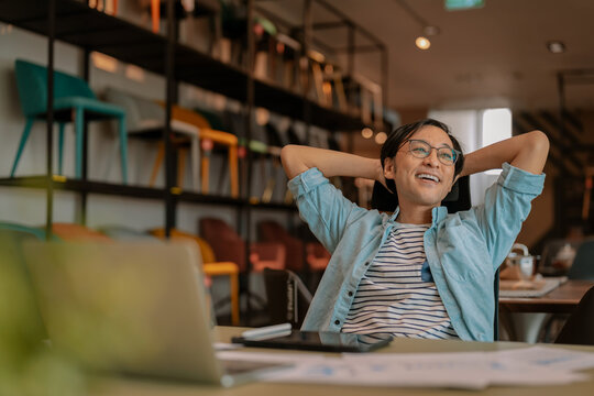 Happy Asian freelancer relaxing comfortably with hands behind head at modern coworking desk, demonstrating career satisfaction, employee wellbeing, contented productivity, work-life balance, and freed