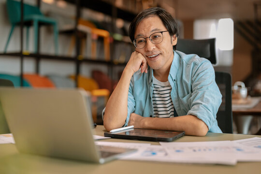 Happy Asian freelancer relaxing comfortably with hands behind head at modern coworking desk, demonstrating career satisfaction, employee wellbeing, contented productivity, work-life balance, and freed