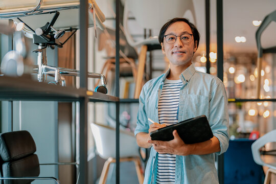 Busy Asian interior designer talking on mobile phone while holding digital tablet in modern furniture showroom, demonstrating professional procurement, client consultation, and inventory coordination.