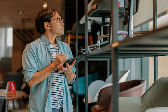 Busy Asian interior designer talking on mobile phone while holding digital tablet in modern furniture showroom, demonstrating professional procurement, client consultation, and inventory coordination.