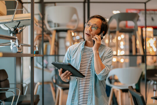Busy Asian interior designer talking on mobile phone while holding digital tablet in modern furniture showroom, demonstrating professional procurement, client consultation, and inventory coordination.
