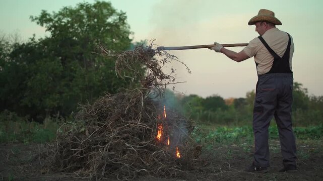 Gardener in straw hat uses pitchfork to tend to burning pile of dry branches in a field, with smoke rising and green trees visible in the background during a warm evening