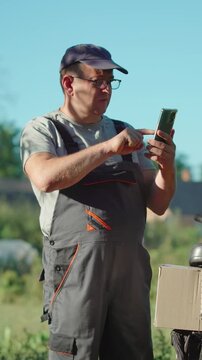 Man in gray overalls and cap interacts with smartphone outdoors, displaying focused expression while standing beside a box and tools in a natural setting with greenery in the background