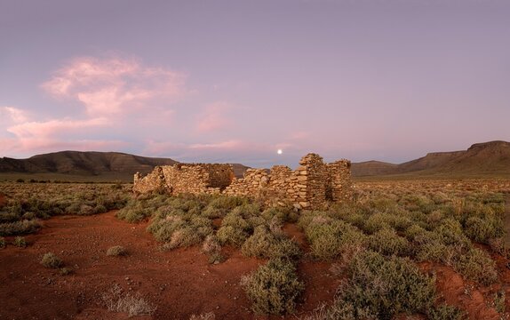 View of stone ruins in a desert landscape with red soil, sparse shrubs, and distant mountains under a twilight sky with a visible moon in South Africa.