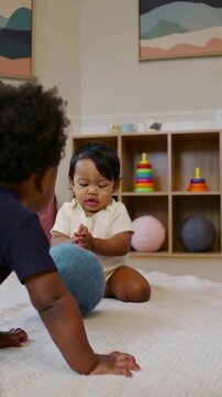 Vertical video: Rolling blue ball moving two babies closer on rug at home, cream onesie palming toy