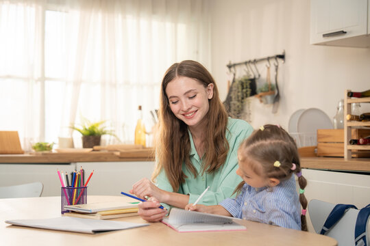 Smiling mother looking at her happy daughter pointing at notebook during home lesson. Young mother encouraging excited little girl student while studying together at table in kitchen.