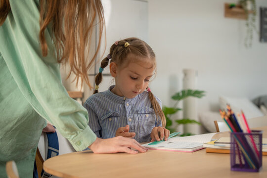 Smiling mother looking at her happy daughter pointing at notebook during home lesson. Young mother encouraging excited little girl student while studying together at table in kitchen.