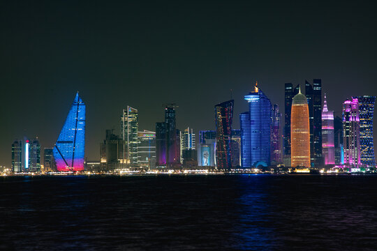 View of the illuminated skyline of Doha at night, featuring modern skyscrapers with vibrant neon lights reflecting over the dark waters of the Persian Gulf in Doha, Doha Municipality, Qatar.