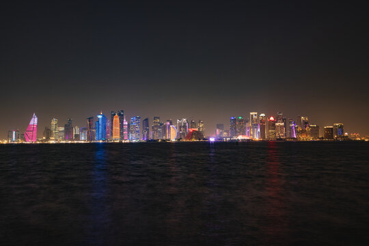 View of the illuminated Doha skyline reflecting on the dark waters of the Persian Gulf at night, featuring modern skyscrapers and architectural landmarks in Doha, Doha Municipality, Qatar.
