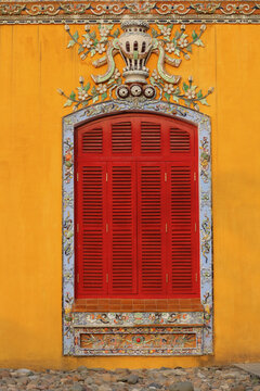 Traditional red wooden louvered shutters on ochre yellow stucco wall with ornate mosaic window frame border and floral stucco relief