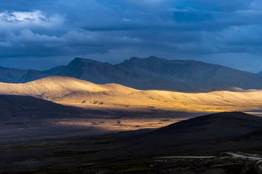 View of Deosai National Park with golden sunlight illuminating the rolling hills against a dramatic dark blue sky in Skardu, Gilgit Baltistan, Pakistan.
