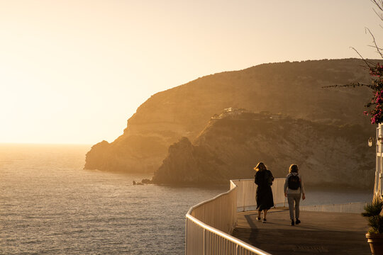 View of two people walking along a coastal path in Ischia with cliffs and the sea illuminated by golden hour light in Ischia, Campania, Italy.