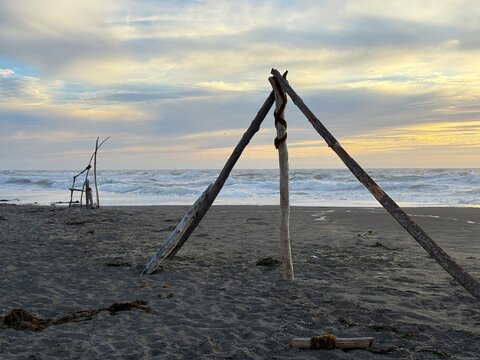 View of driftwood structures on a sandy beach with ocean waves under a soft sunset sky in Fort Bragg, California, United States.