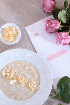 Oatmeal with coconut chips in white plate. Small bowl with coconut. Pink roses and card on beige background. Top view healthy breakfast. Flat lay