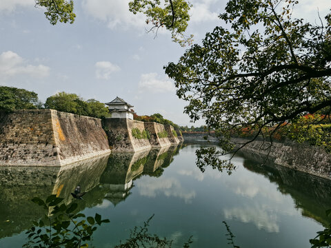 View of the historic stone walls and moat surrounding Osaka Castle with lush trees and a white turret reflecting in the calm water in Osaka, Osaka, Japan.