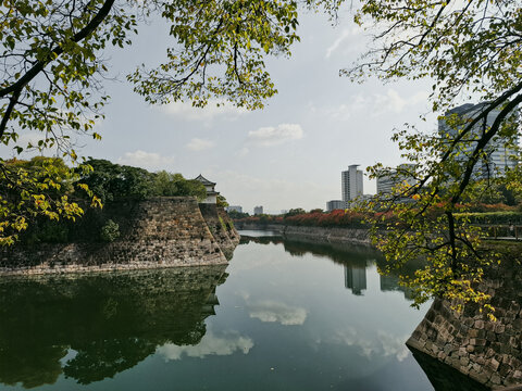 View of the historic stone walls and moat surrounding Osaka Castle, framed by autumn foliage with modern city buildings in the background in Osaka, Osaka, Japan.