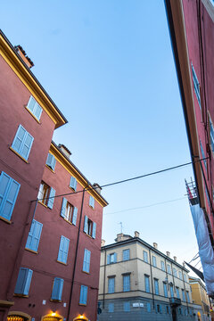 Aerial view of historic buildings with red and yellow facades and blue shutters under a clear blue sky in Modena, Emilia-Romagna, Italy.