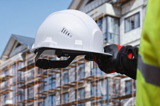 Construction worker holding a white safety helmet and wearing protective gloves in front of a building under construction. Residential development, construction work and occupational safety equipment.