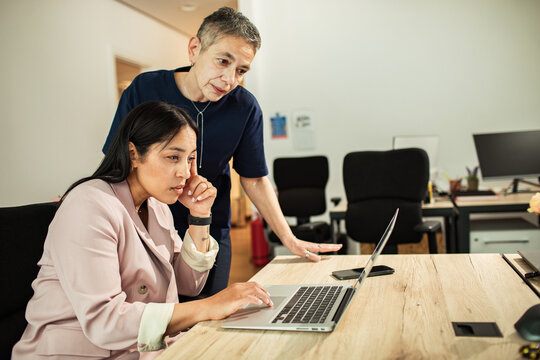 Manager guiding colleague at laptop in modern office