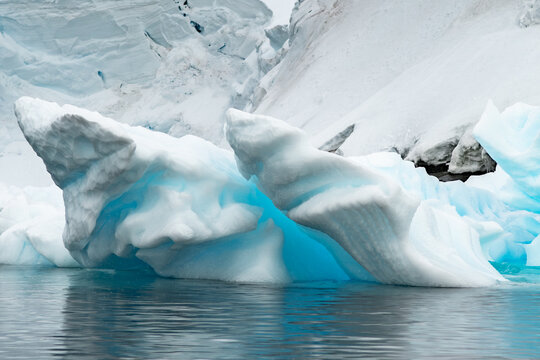 View of a large, bright blue iceberg floating in the calm waters with massive snow-covered glaciers and icy peaks rising in the background Antarctica.