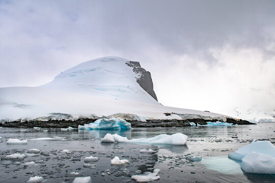 View of a snow-covered mountain peak rising above icy waters filled with floating icebergs in Antarctica.