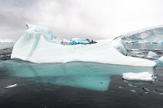 View of a large, jagged iceberg floating in dark, icy water with a cloudy sky in the background in Antarctica.