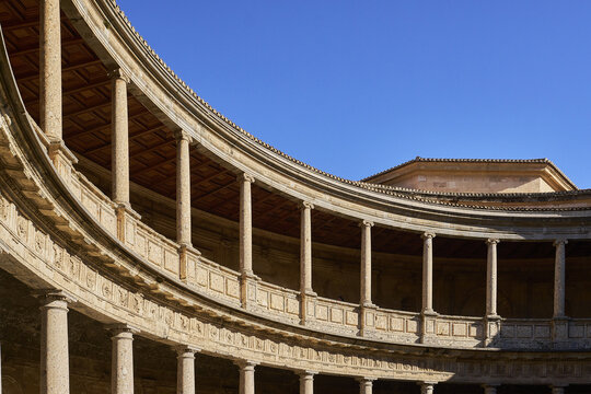 View of the circular courtyard of the Palace of Charles V with its two-story colonnade, stone columns, and ornate carvings under a clear blue sky in Andalusia, Spain.