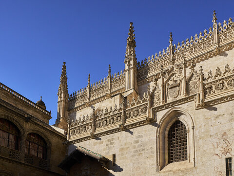 View of the ornate Gothic stone facade of the Royal Chapel of Granada with intricate carvings, pinnacles, and arched windows under a clear blue sky in Andalusia, Spain.