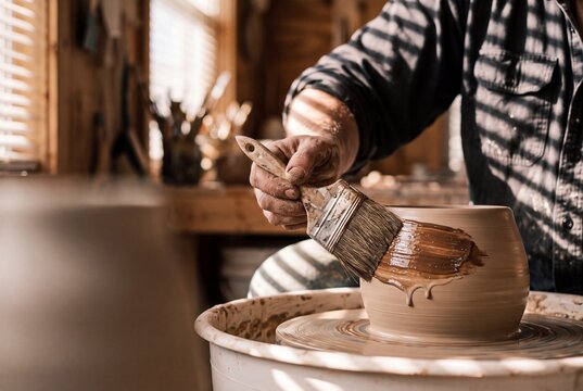 Close up of a skilled craftsman applying liquid glaze with a brush to a fresh clay pot on a spinning pottery wheel in a warm sunlit workshop