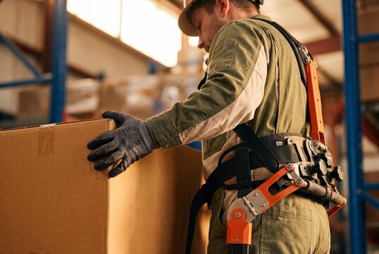 Male warehouse worker wearing modern supportive exoskeleton suit lifting heavy cardboard box in logistics distribution center
