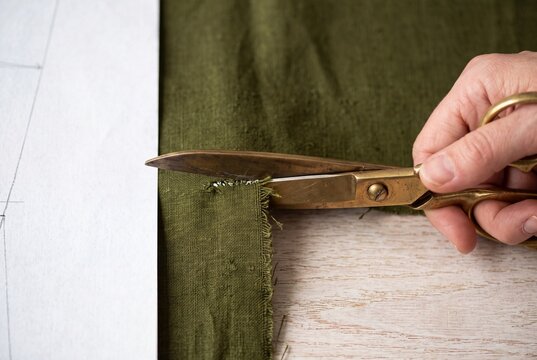 A seamstress uses vintage brass shears to cut green linen fabric along a paper sewing pattern on a wooden table