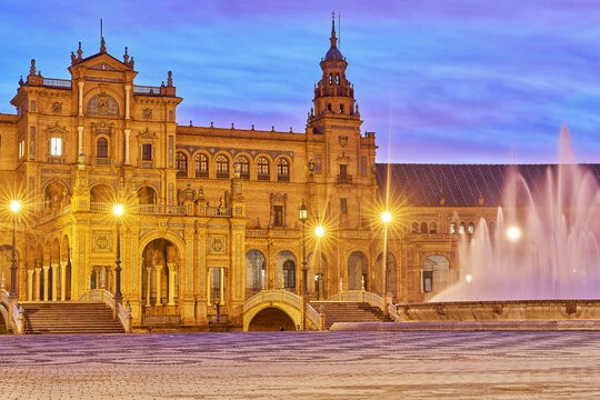 View of the illuminated Plaza de Espana with its ornate architecture, central fountain, and glowing street lamps at dusk in Seville, Andalusia, Spain.