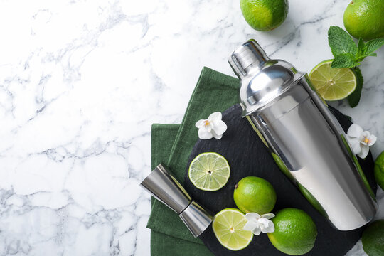 Shaker, limes, orchid flowers, mint and jigger on white marble table, flat lay. Space for text