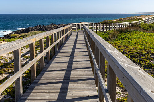 View of a wooden boardwalk winding along the rugged coastline with rocky shores and green vegetation overlooking the ocean in Sines, Setubal, Portugal.